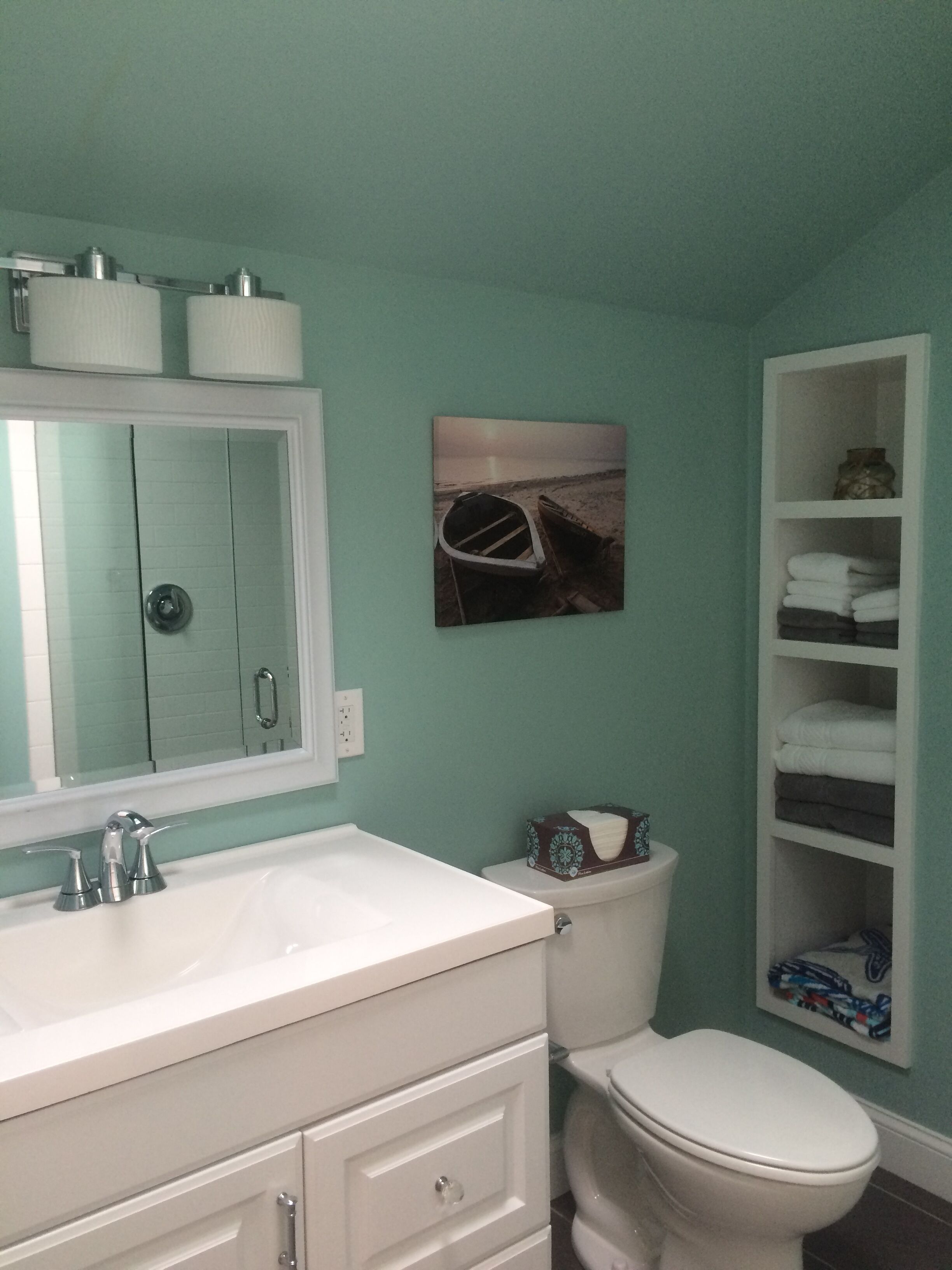 Un bagno con lavandino bianco, specchio e WC. Si nota uno scaffale con asciugamani e un quadro raffigurante una barca appeso al muro.