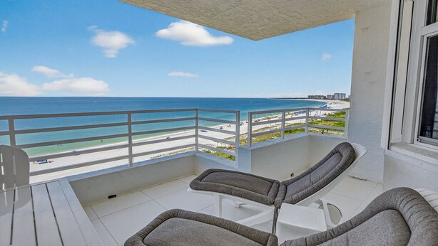 Ein Balkon mit zwei gepolsterten Stühlen und einem Tisch, mit Blick auf einen Strand und eine freie Sicht auf das Meer und entfernte Gebäude.