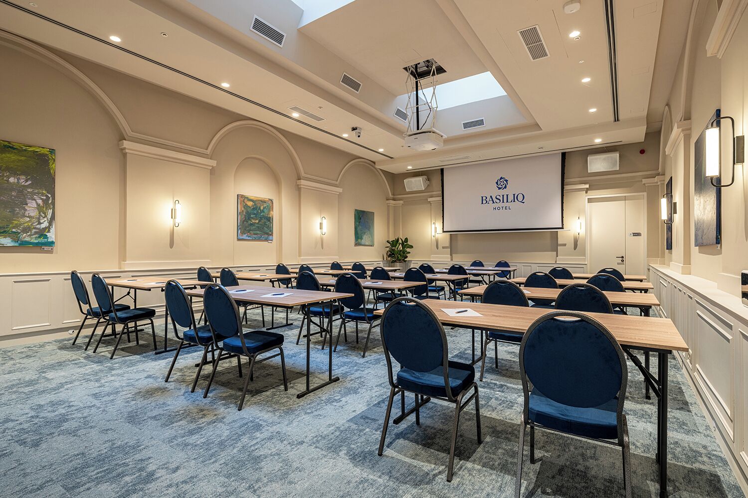 A conference room with a large screen displaying 'BASILIQ HOTEL' branding, multiple tables with chairs, and a carpeted floor.