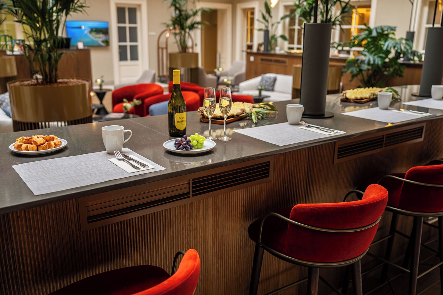 Un coin bar avec un comptoir en bois, des chaises rouges, une bouteille de vin et une assiette de raisins.