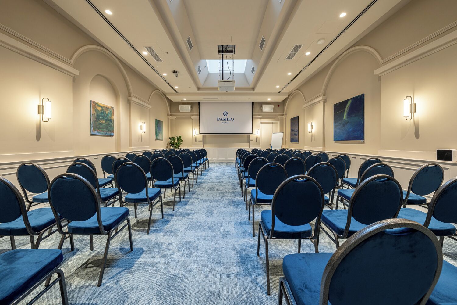 Un salón de conferencias con filas de sillas azules, una pantalla de proyector y un letrero que dice 'BASILIQ Hotel'.