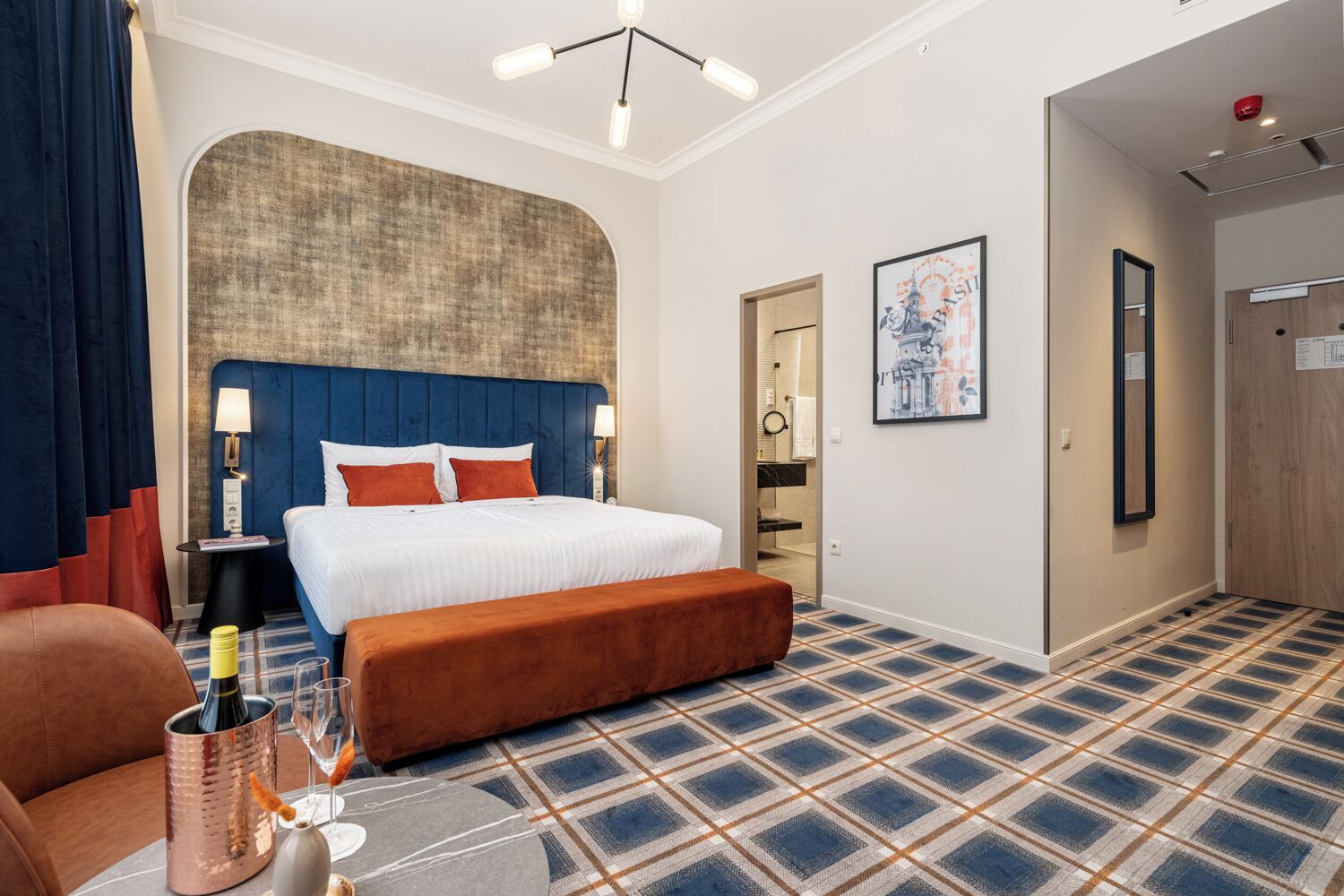 A hotel room with a large bed, a bench, a small table with a bottle and glasses, a wall-mounted lamp, and a door leading to a bathroom.
