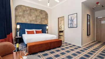 A hotel room with a large bed, a bench, a small table with a bottle and glasses, a wall-mounted lamp, and a door leading to a bathroom.
