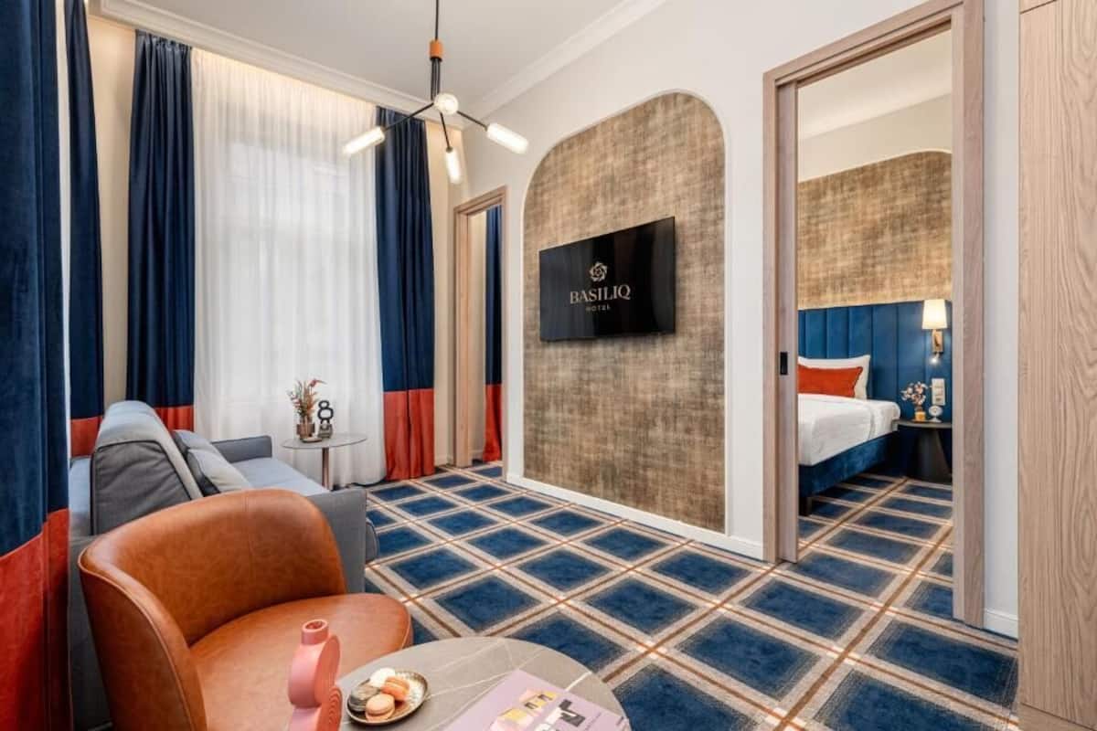 Una habitación de hotel con alfombra azul y beige a cuadros, un sofá, una mesita con una revista y un objeto rosa, y una cama con cabecero azul.