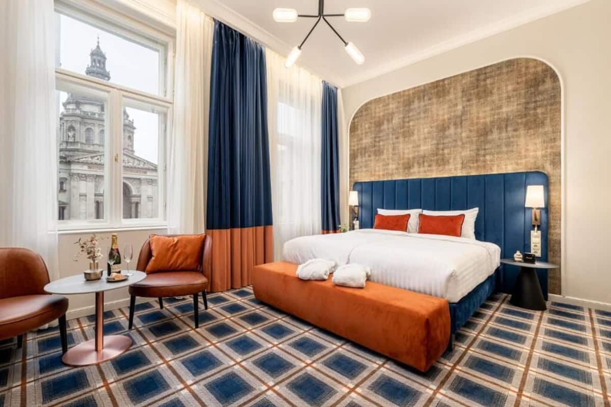 Habitación de hotel con una cama grande, una mesita con una botella y vasos, un sofá y vistas a un edificio histórico.