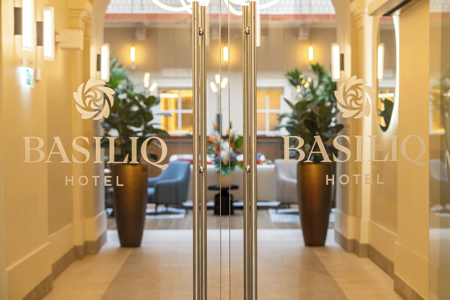 Entrada de hotel con diseño moderno, destacando un acceso de vidrio, una zona de recepción con asientos y plantas en macetas.