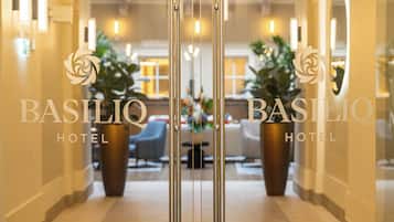 Entrada de hotel con diseño moderno, destacando un acceso de vidrio, una zona de recepción con asientos y plantas en macetas.