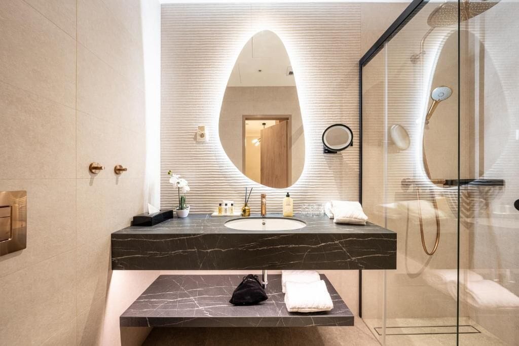 Un baño moderno con un espejo ovalado grande, encimera de mármol, cabina de ducha de cristal y un banco con toallas.