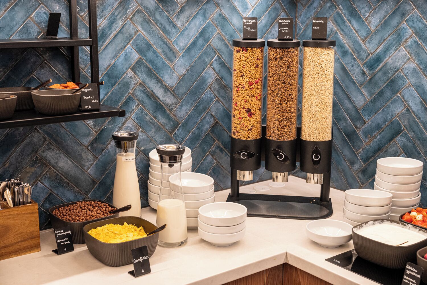 A buffet setup with various food items and containers, including a milk dispenser and a cereal bowl.