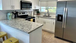 Una cocina con armarios blancos, un refrigerador de acero inoxidable, un microondas y una estufa. Hay una ventana que ofrece vistas a zonas verdes.