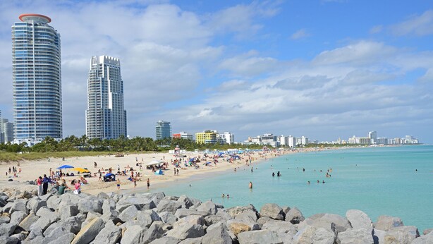 Una playa con rocas grandes al frente, una orilla arenosa y edificios altos al fondo.