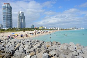 A beach with large rocks in the foreground, a sandy shore, and tall buildings in the background. - Stay like a local in this 1BR/1BA near Brickell. <br>Central location with parking, private patio, and fast WiFi. <br>Minutes to restaurants, cafés, nightlife, and everything Miami offers. (Miami)