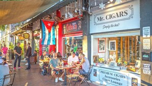 Escena callejera con mesas al aire libre en un café y una tienda de cigarros.