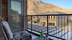 Ein Balkon mit einem Korbstuhl, einer Flasche und einem Glas. Der Balkon bietet Blick auf einen Parkplatz und ein Gebäude vor einer hügeligen Landschaft.