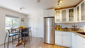 Een moderne keuken met witte kastjes, een roestvrijstalen koelkast, een eethoek met een ronde tafel en stoelen, en een raam dat natuurlijk licht binnenlaat.