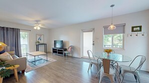 A spacious living area with a sofa, TV, glass table, chairs, and a ceiling fan.