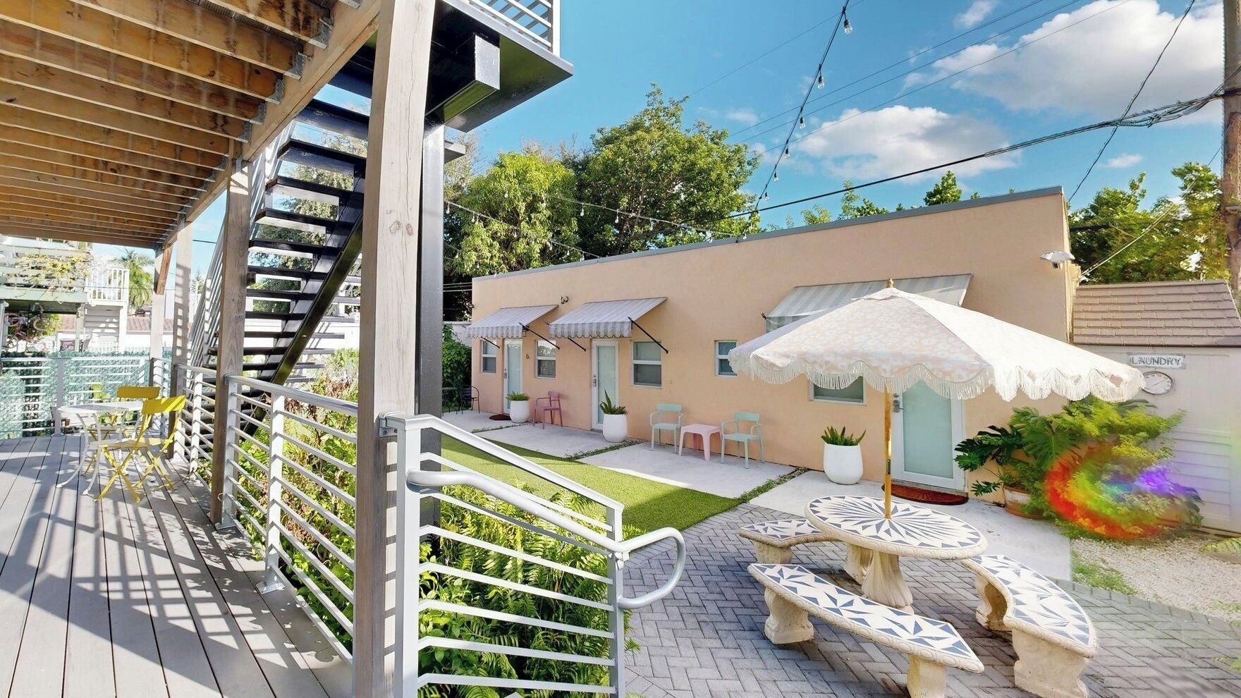 A modern outdoor patio with a wooden deck, white outdoor furniture, and a small building with a beige facade.