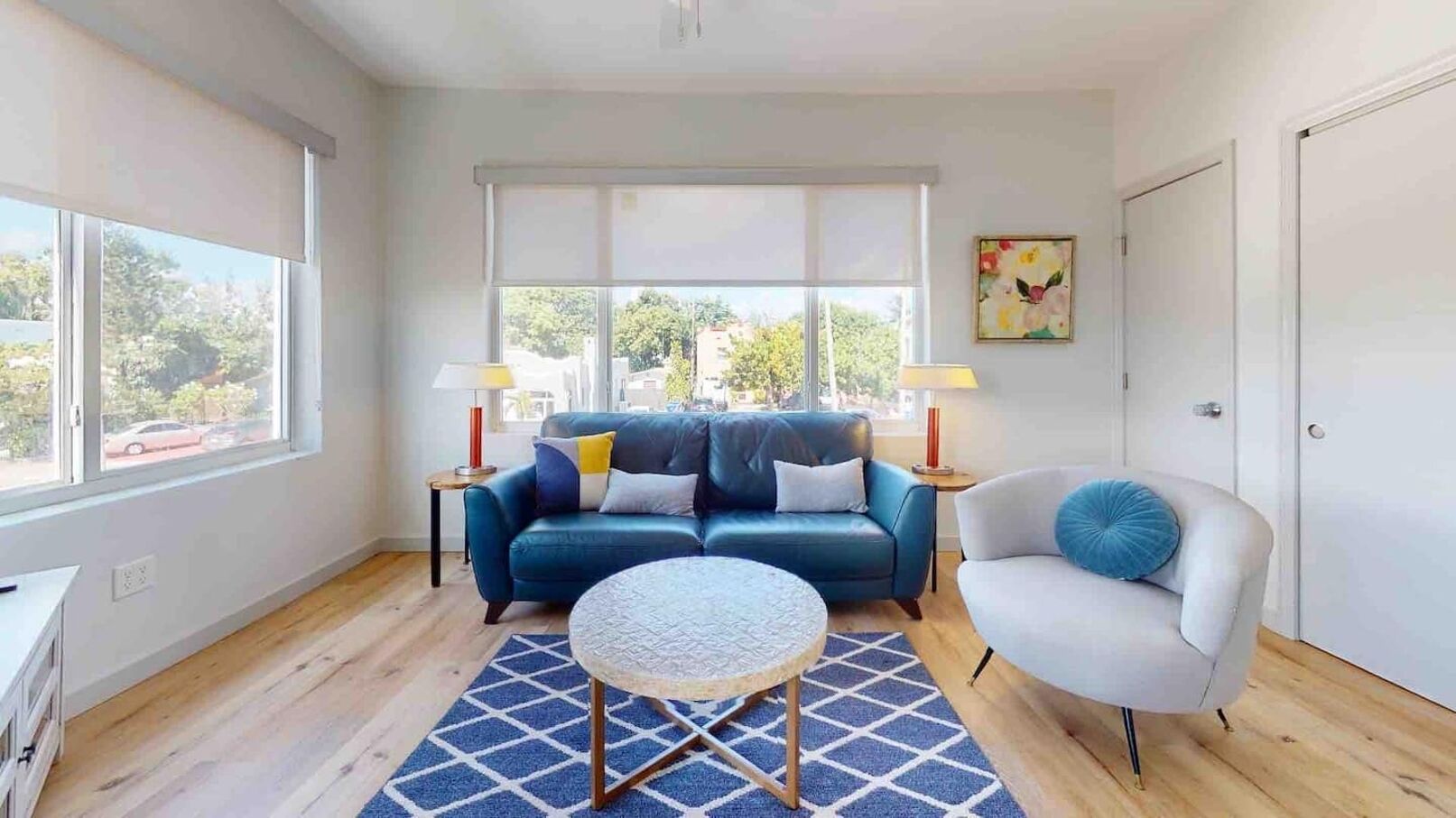 Een moderne woonkamer met een blauwe bank, een ronde salontafel en een witte fauteuil. Er staan twee bijzettafels met lampen en er hangt een schilderij aan de muur.