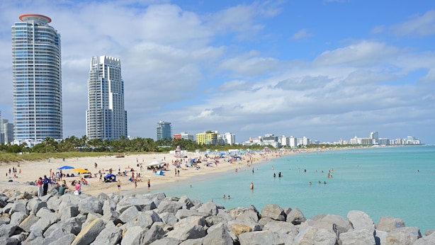 Une plage avec de gros rochers au premier plan, une étendue de sable et de grands immeubles en arrière-plan.
