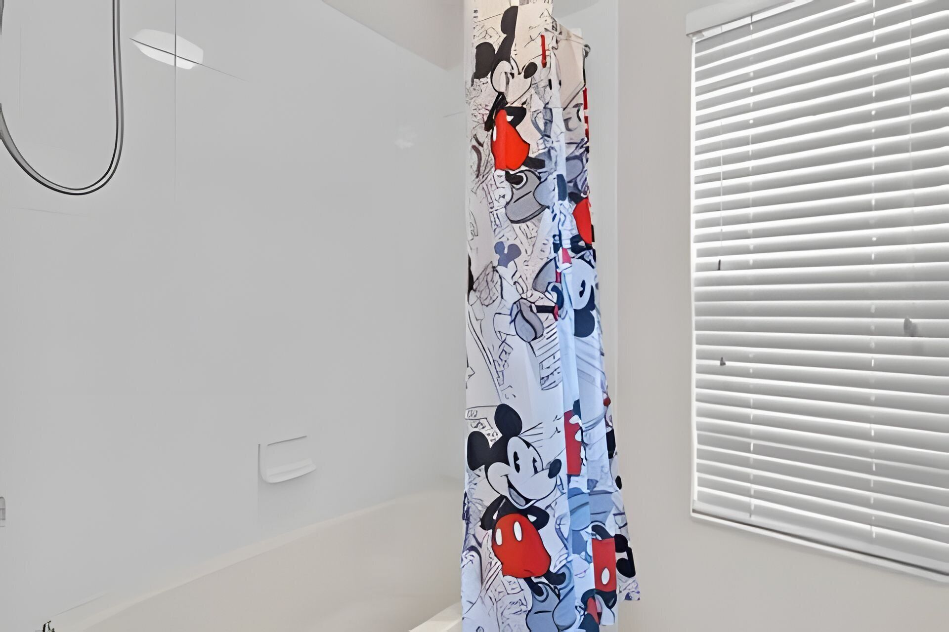 A bathroom with a Mickey Mouse themed shower curtain, white walls, and a window with blinds.
