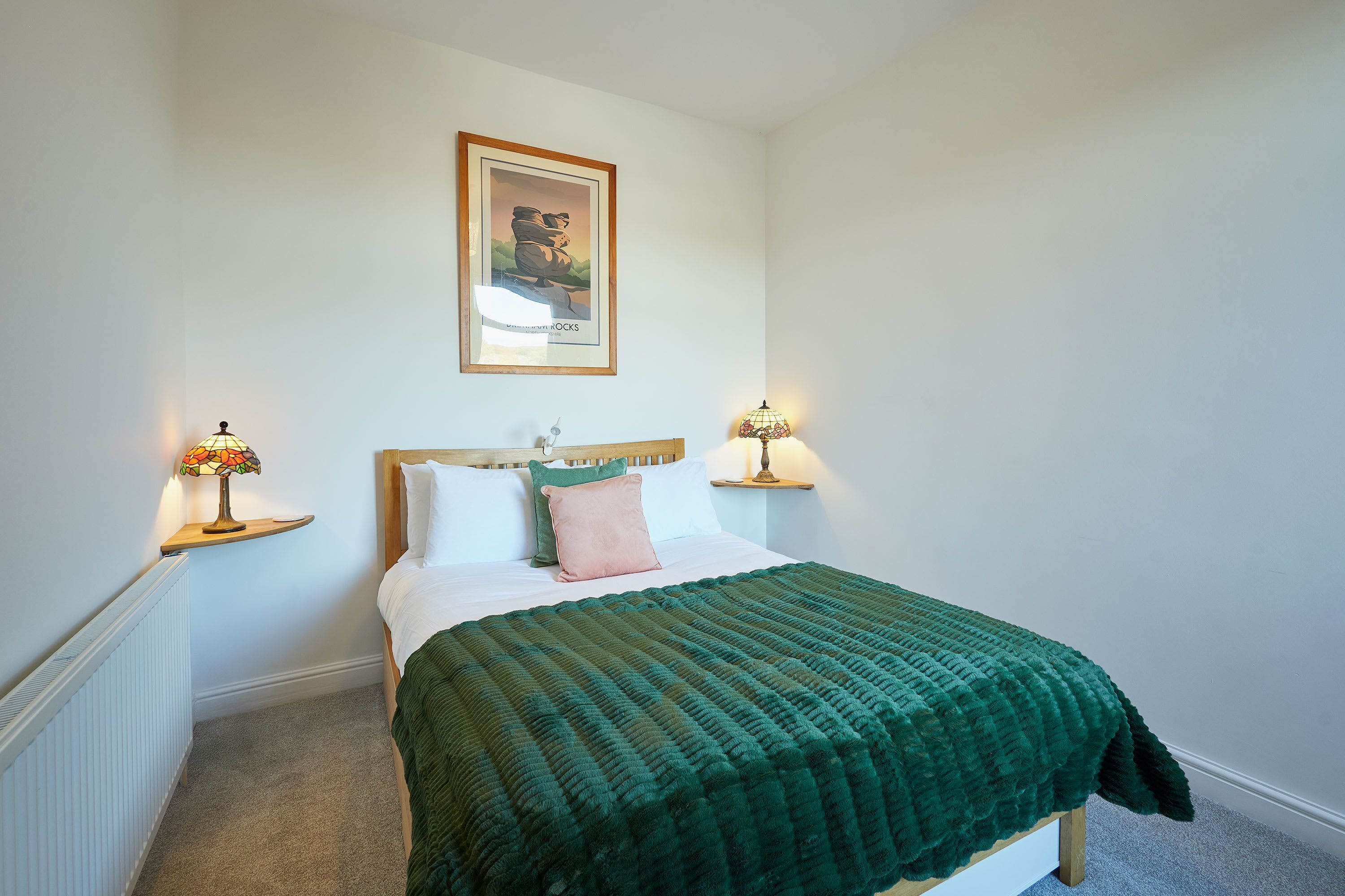 Un letto rifatto con cura, con una coperta verde, due abat-jour, un quadro incorniciato sopra il letto e una piccola mensola sul comodino.