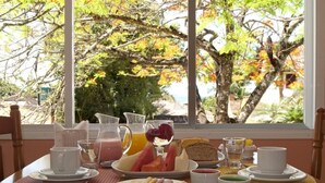 A breakfast table with plates of food, cups, and a pitcher of juice, set against a window with a view of trees and a beach. - Azul da Cor do Mar (São Sebastião)