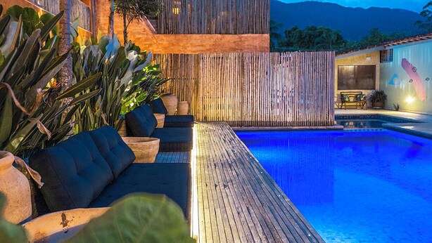 A modern pool area with a wooden deck, outdoor seating, and a view of the mountains. - Azul da Cor do Mar (São Sebastião)