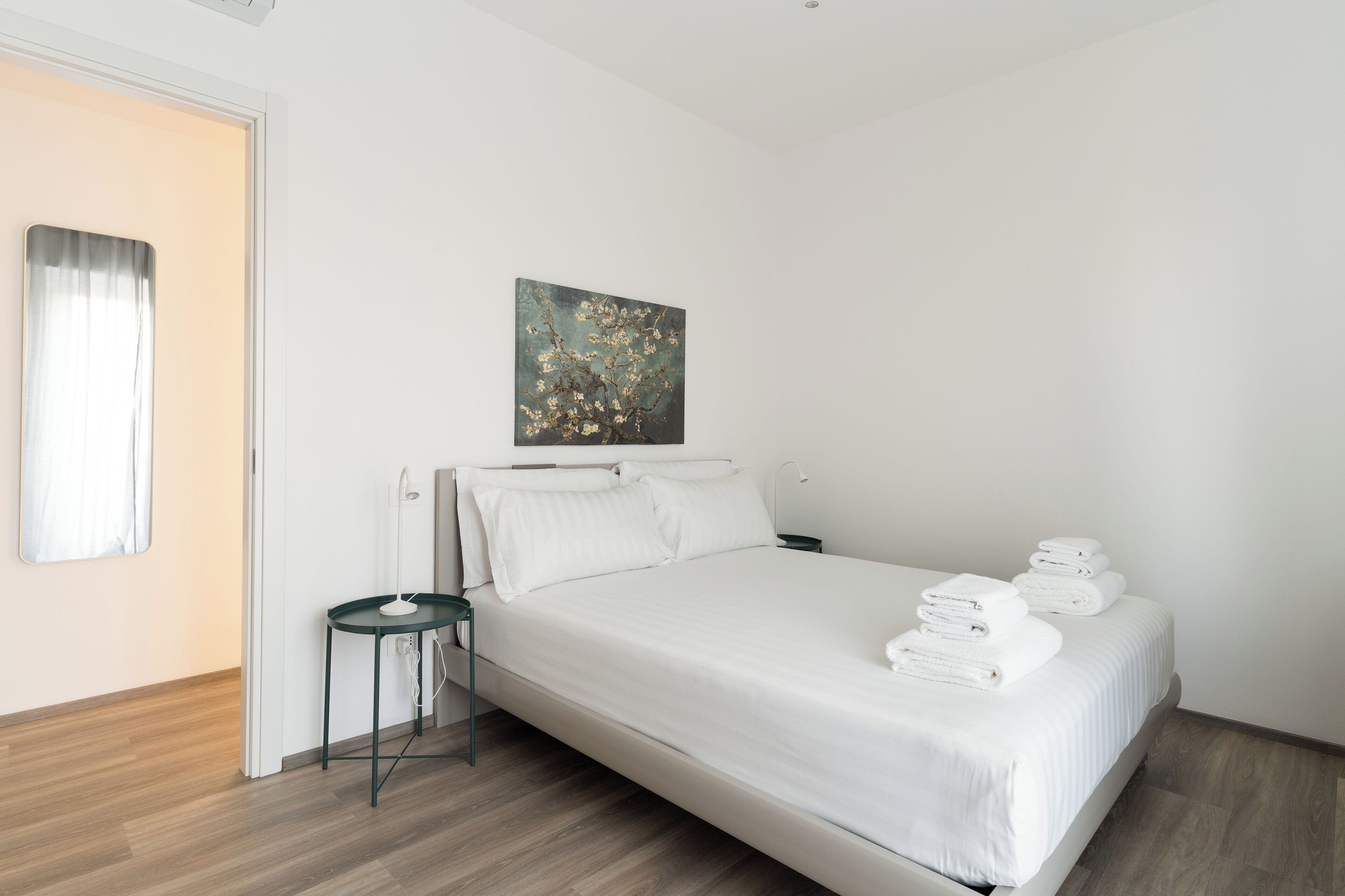 A neatly made bed with white linens and towels, a small bedside table, a wall-mounted painting, and a wooden floor.