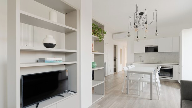 A modern living room with a flat-screen TV, white shelving, a dining area with a table and chairs, and a kitchen with white cabinets and a built-in oven. - Brera Apartments Navigli (Milan)