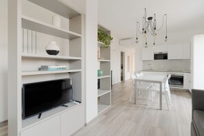 Una sala moderna con televisor de pantalla plana, estanterías blancas, un comedor con mesa y sillas, y una cocina con armarios blancos y horno empotrado.