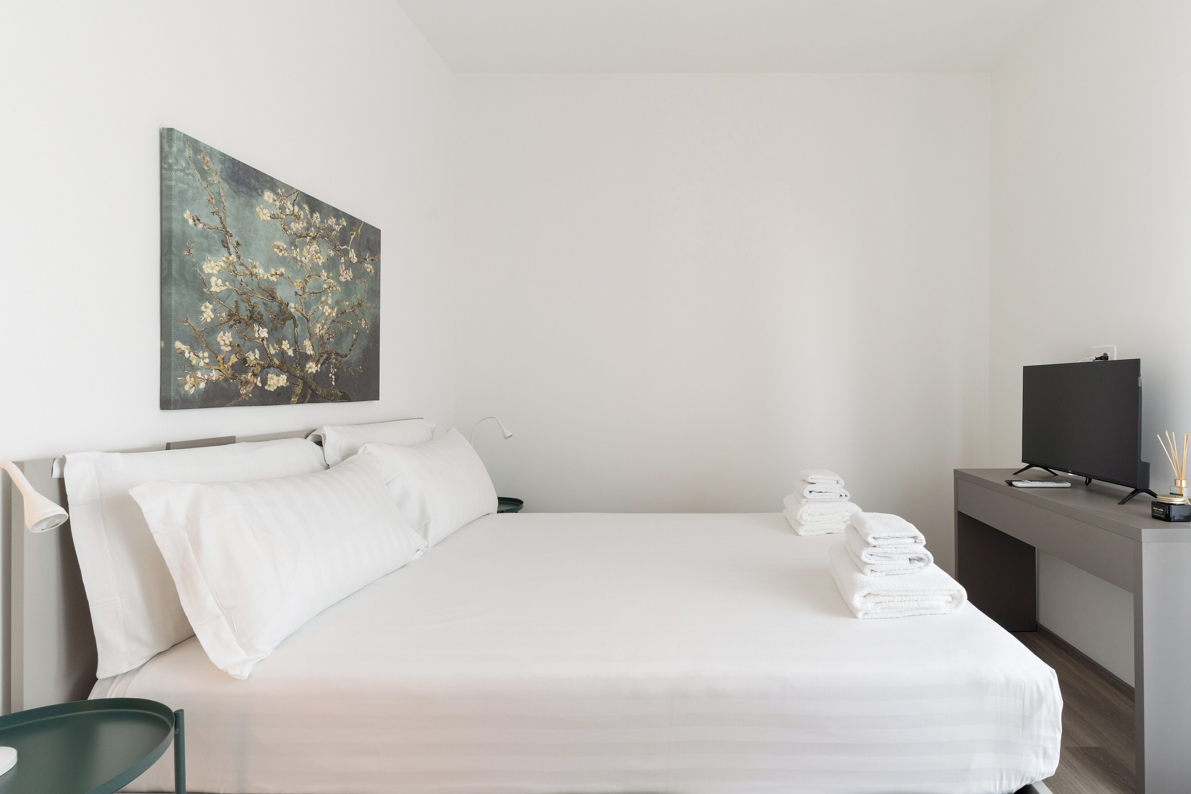 Ett modernt hotellrum med en snyggt bäddad säng, en tavla på väggen, en platt-TV och ett skrivbord med en lampa och en flaska med doftljus.