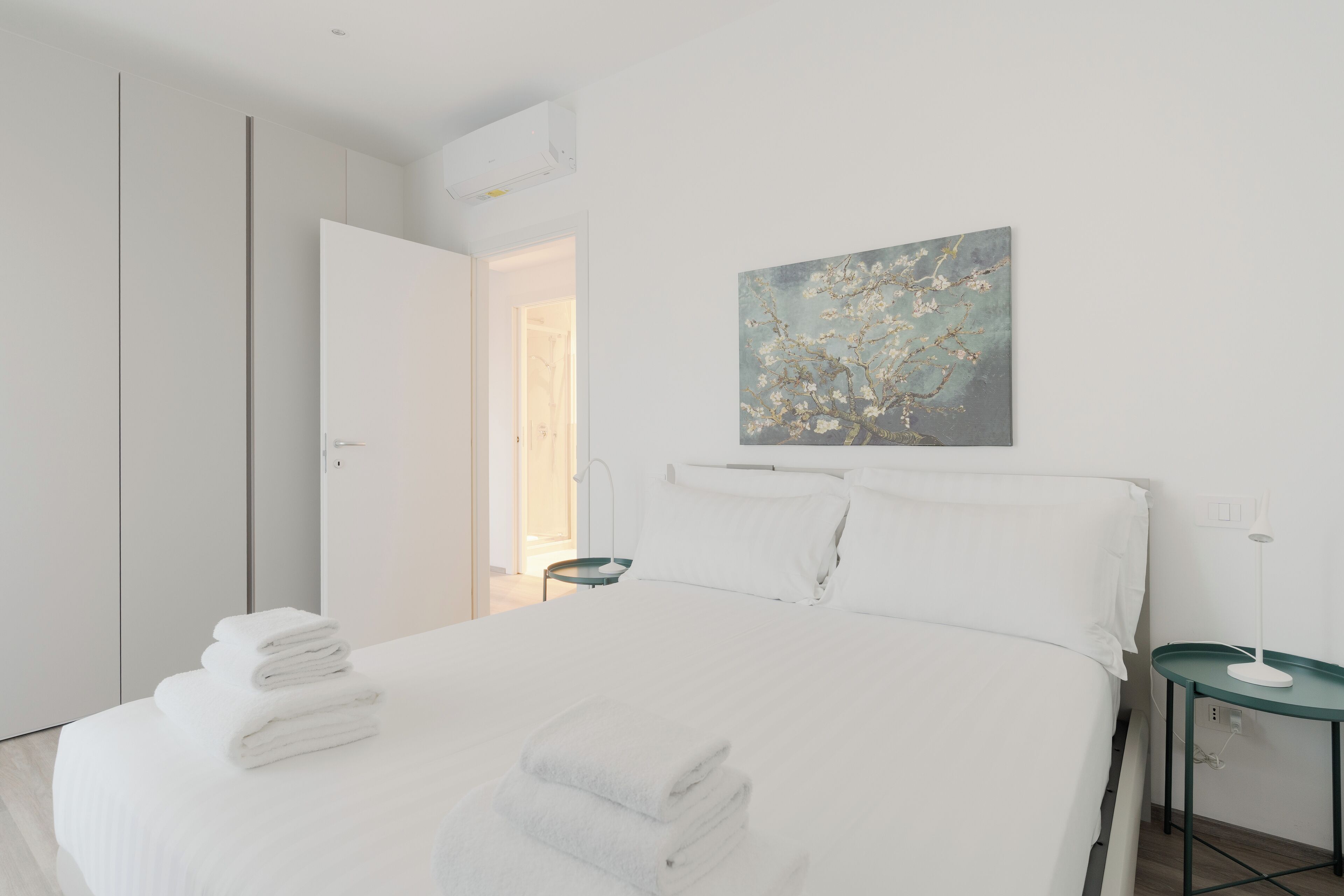 A modern bedroom with a neatly made bed, a wall-mounted artwork, and a small bedside table with a lamp.