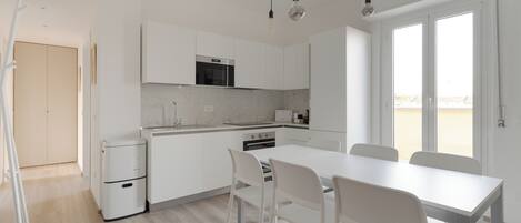 Una cocina moderna con armarios blancos, horno empotrado y comedor con mesa y sillas blancas.