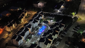 Vista aérea de una zona residencial con varios edificios blancos, una piscina con iluminación azul y un estacionamiento.