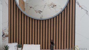 Une salle de bain avec un mur recouvert de lambris en bois, un miroir rond, un pare-feu en marbre, un lavabo blanc, un robinet noir et une petite plante en pot.
