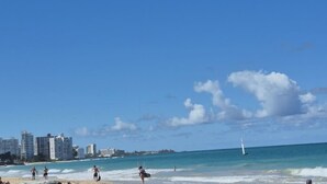 Ein Strand mit Gebäuden im Hintergrund, einem klaren Himmel und einem Segelboot auf dem Wasser.