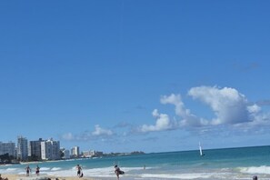 A beach with buildings in the background, a clear sky, and a sailboat in the water. - Condado Condo Retreat | Private Patio (San Juan)