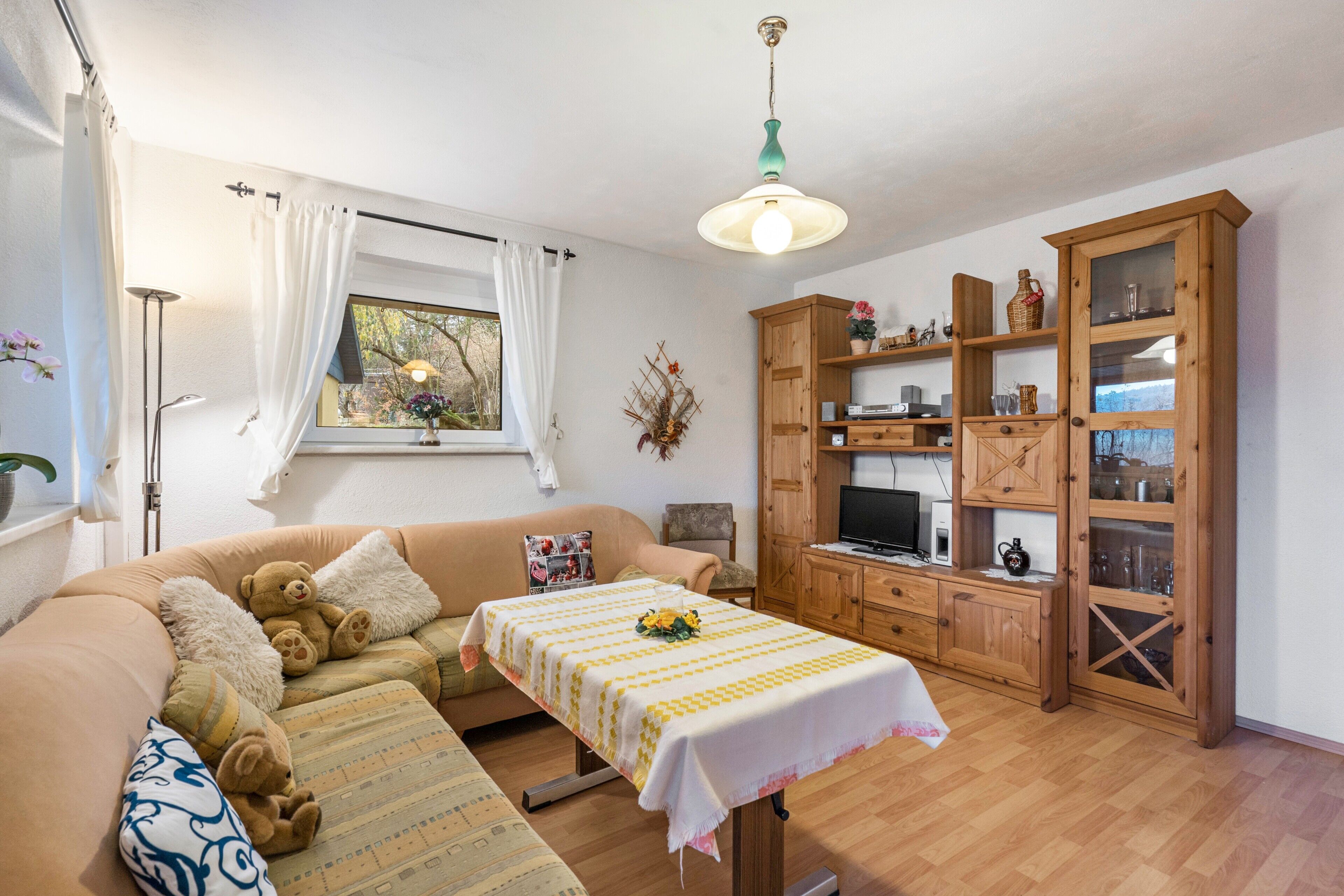 Una sala de estar acogedora con un sofá, una mesa con mantel, un aparador de madera con puertas de cristal y una lámpara de techo.