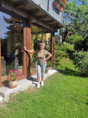 A woman standing outside a modern building with large glass doors, a wooden porch, and a garden with potted plants. - Heidi’s Holiday Apartment with Mountain View, Private Garden & Wi-Fi (Rosenthal-Bielatal)