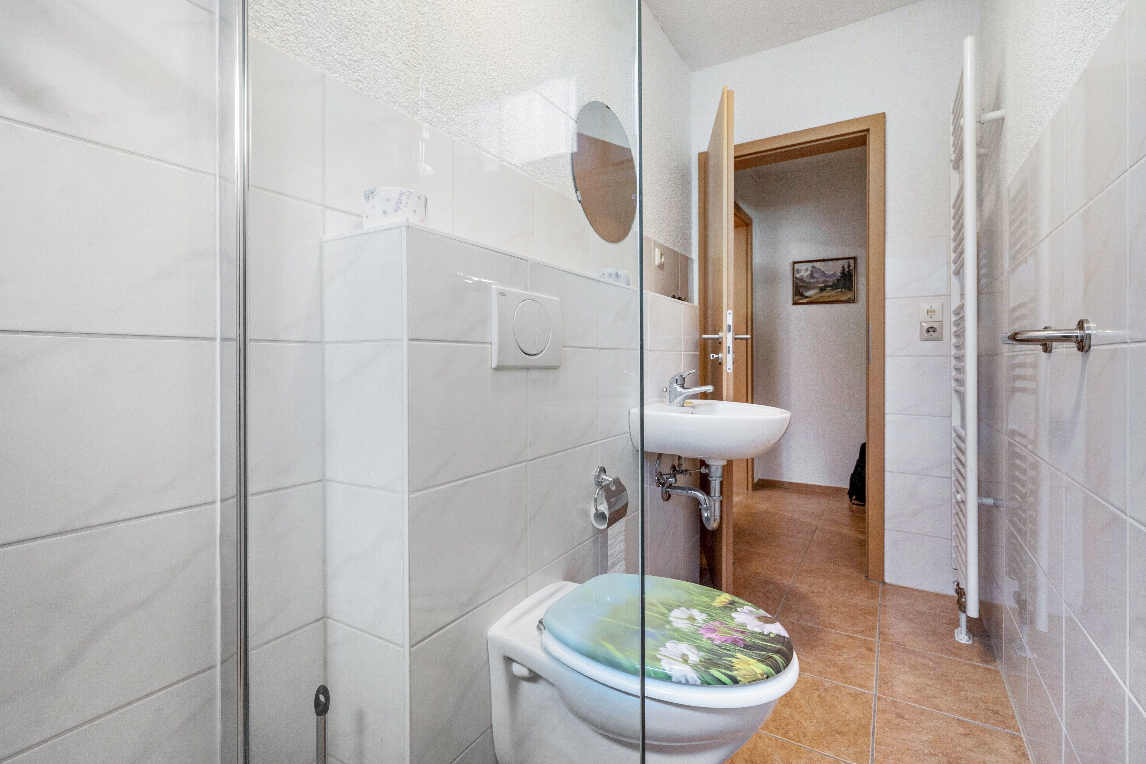 A bathroom with a glass shower enclosure, a toilet with a floral seat, a sink, and a towel rack.