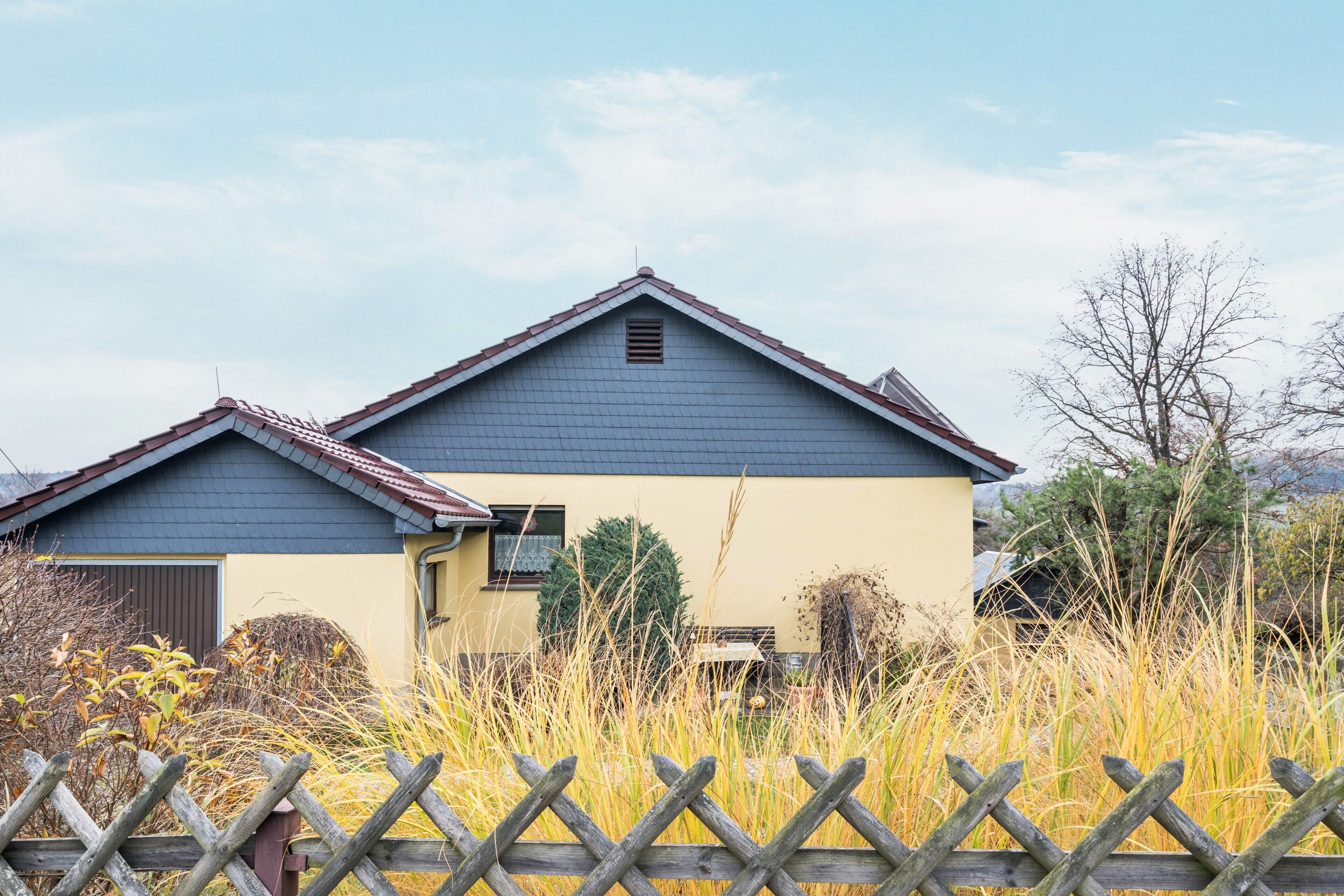 Een huis met twee verdiepingen, met een blauw dak en een geel gevel, omringd door hoog gras en een houten hek.