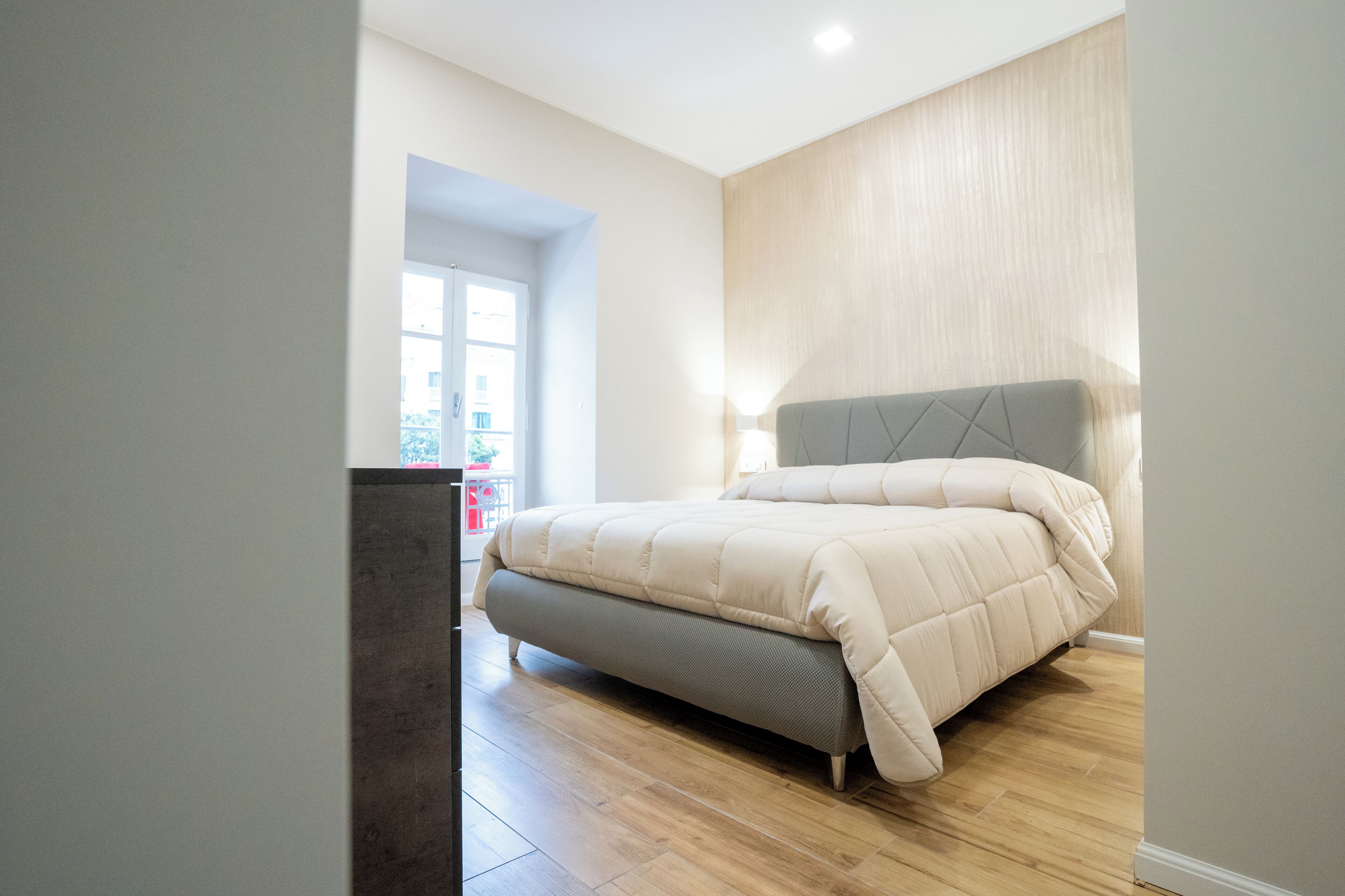 A bedroom with a bed, wooden flooring, and a view through an open door.