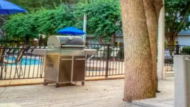 Een houten terras met een grill en barbecue, een zwembad op de achtergrond, en een boom.