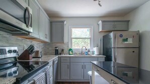 A modern kitchen with grey cabinets, a stainless steel refrigerator, a built-in oven, and a window above the sink. - Charming 3BR Home w/ Fenced Yard Near DT Raleigh (Raleigh)