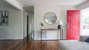 A room with hardwood flooring, a grey sofa, a red door, a round mirror, a small table with a lamp, and framed pictures on the wall. - Charming 3BR Home w/ Fenced Yard Near DT Raleigh (Raleigh)
