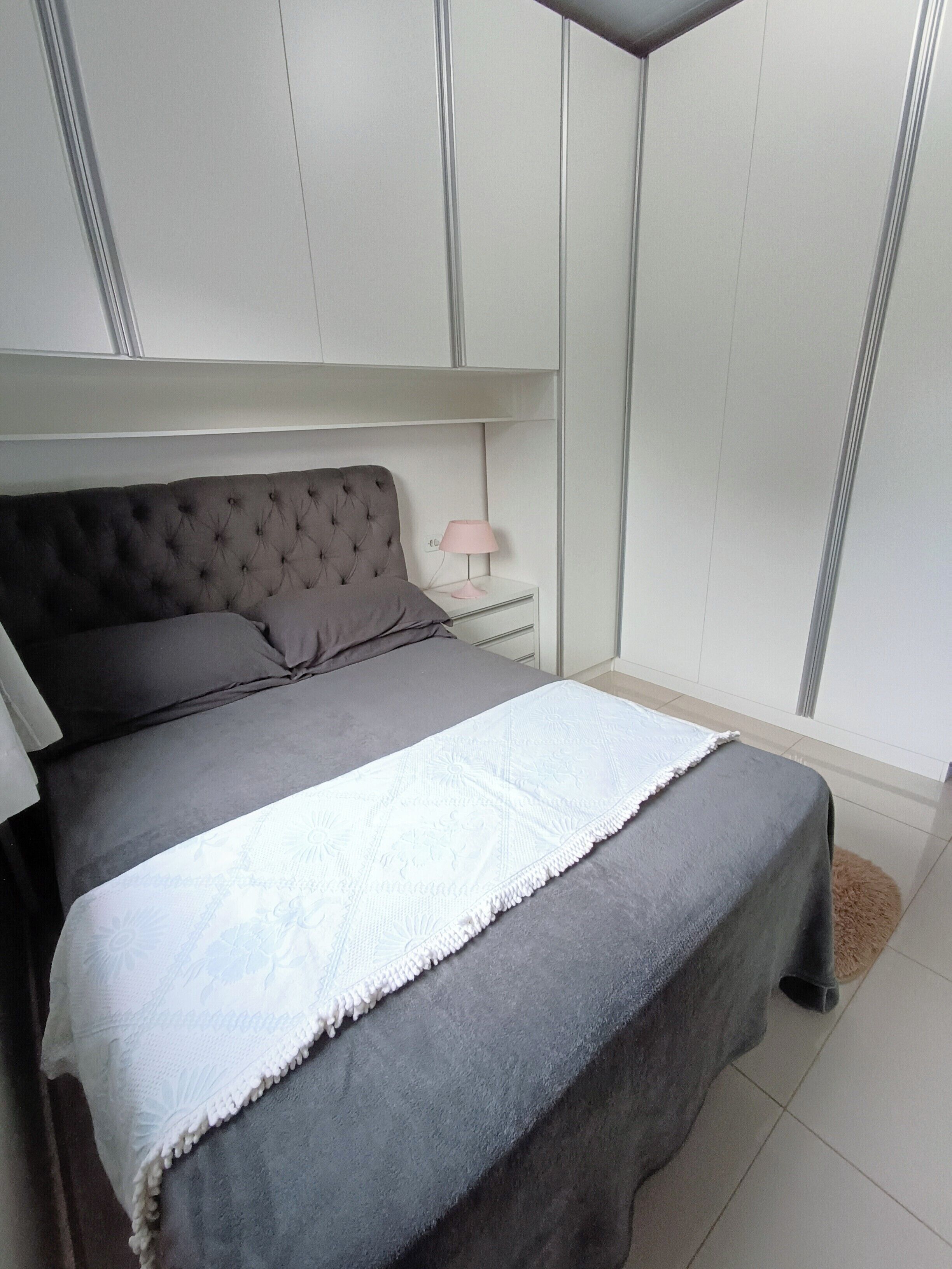 A bedroom with a grey upholstered bed, a white bedside table with a pink lamp, and a wardrobe with vertical handles.