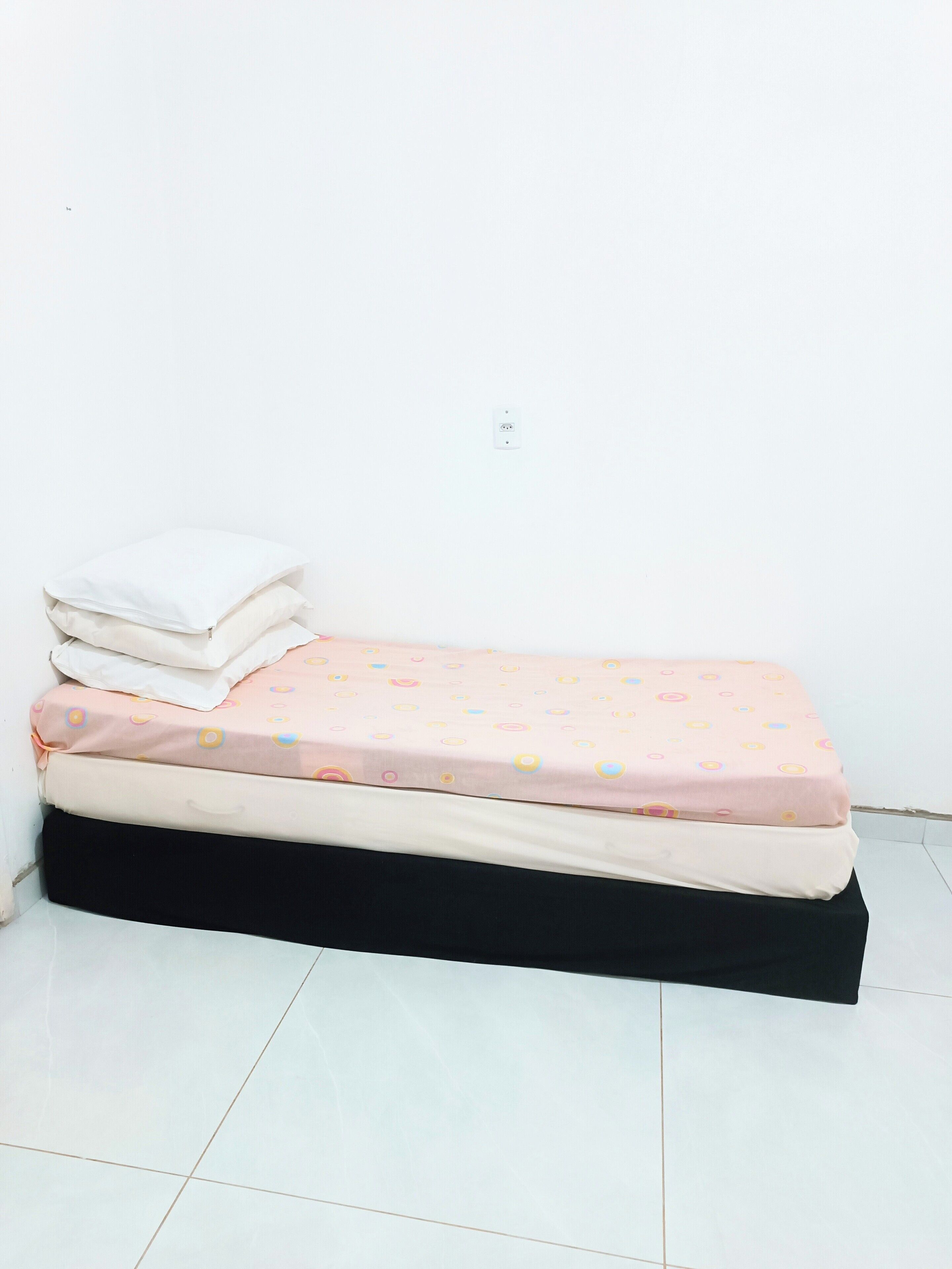 Un piccolo letto con materasso rosa e cuscini bianchi, su uno sfondo di parete bianca.