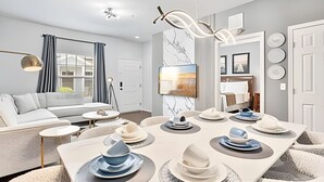 A modern living room with a sofa, a dining table set with plates and bowls, a TV mounted on the wall, and a large window with curtains. - 3BR Lucaya Village Home Near Disney (Kissimmee)