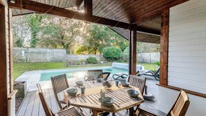Eine überdachte Terrasse im Freien mit einem Holztisch und -stühlen, einem Pool und Blick auf den Garten.