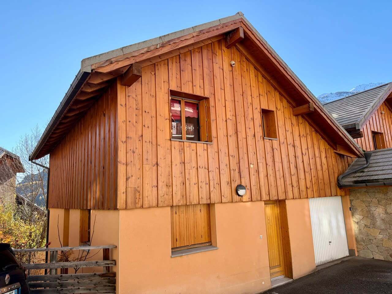Un chalet en bois avec un toit en pente, une petite fenêtre ornée d’un rideau rouge et un mur de pierres attenant au bâtiment.